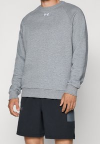 Sweat-shirt gris avec un col rond et des poignets côtelés. Présente un petit logo blanc sur la poitrine. Associé à un short noir, montrant un design épuré.