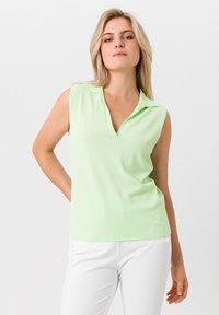 Lichtgroene mouwloze polo met een V-hals, zachte textuur en een relaxte pasvorm, gecombineerd met witte broeken. Eenvoudig en modern ontwerp.