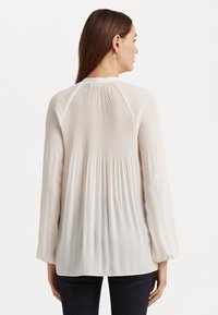 Krämfärgad blus med långa, puffiga ärmar, har vertikala veck och en smockad bakdel. Tyget verkar lätt och texturerat.