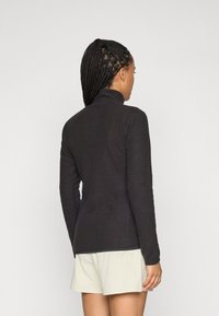 Vrouw met lang gevlochten haar, gekleed in een zwart, gestructureerd long-sleeve top en lichtgekleurde shorts, die met haar rug naar de camera staat tegen een effen achtergrond.