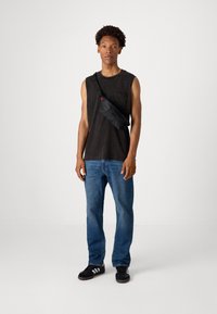 Camiseta sin mangas negra, jeans de mezclilla azul y zapatillas negras con rayas blancas. Se lleva una riñonera negra cruzada. Fondo limpio.