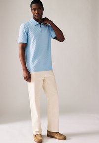 Svetle modrá polokošile s krátkým rukávem a texturovaným vzorem, zkombinovaná s krémovými kalhotami a hnědými nazouvacími botami.