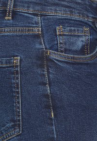 Jeans de ganga azul escuro com costura laranja, apresentando um bolso traseiro, uma textura suave e um padrão de trama visível.