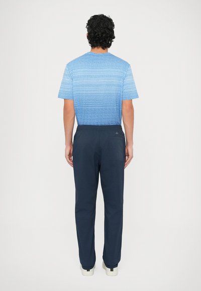 Koszula w niebieskie paski z krótkim rękawem, widok od tyłu, w zestawieniu z granatowymi spodniami. Spodnie mają boczną kieszeń; buty są białe z kolorowymi akcentami.