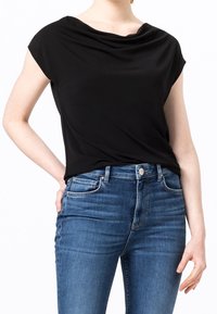 Kvinna iklädd en svart kortärmad topp med draperad hals som är stoppad i högmidjade blå jeans, med vänsterhanden vilande på höften.