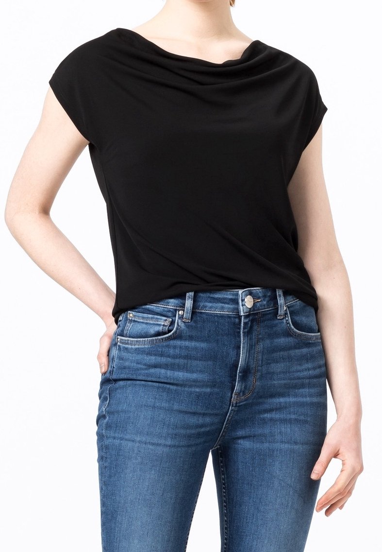 Kvinna iklädd en svart kortärmad topp med draperad hals som är stoppad i högmidjade blå jeans, med vänsterhanden vilande på höften.