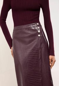 Burgundska rebrasta pletenina zgoraj v kombinaciji z sijočo, teksturirano midi krilom iz umetnega usnja z vzorcem krokodila in štirimi okrasnimi srebrnimi gumbi.