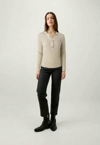 Beige gerippter Pullover mit Henley-Ausschnitt und Knopfdetails, kombiniert mit dunkler Jeans und schwarzen Stiefeletten. Einfaches und tailliertes Design.