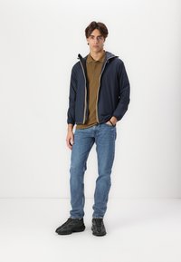 Navy lightweight jacket with contrasting yellow zipper, brown polo shirt, blue jeans, and black athletic shoes. Minimal design with practical features.