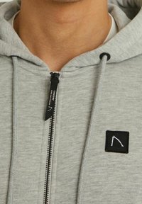 Primer plano de una persona que lleva un sudadera con capucha de cremallera en gris claro, con un parche rectangular de logo negro y una lengüeta de cremallera negra.