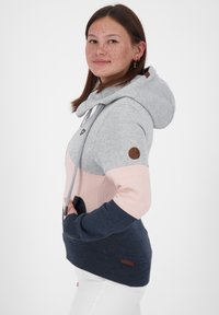 Giovane donna con lentiggini che indossa un hoodie grigio, rosa e blu navy a blocchi di colore con pantaloni bianchi, mani in tasca, rivolta di lato con un leggero sorriso.