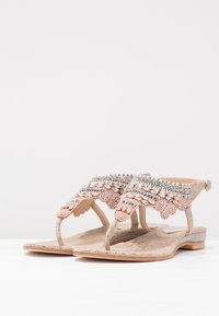 Beżowe płaskie sandały z misternymi różowymi, srebrnymi i kryształowymi ozdobami oraz paskami na kostkę na białej powierzchni.
