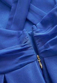 Sininen kangasvaate, jossa on poimutettu yksityiskohta; siinä on kullanvärinen vetoketju, sileä pinta ja monimutkainen poimutus sulkemiskohdan lähellä.