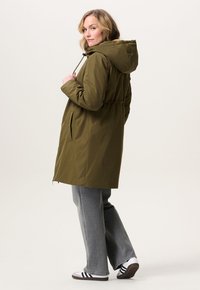 Vrouw in een olijfgroene capuchonparka, grijze wijde jeans en witte sneakers met zwarte strepen, kijkend over haar schouder.