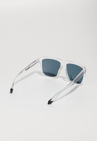 Lunettes de soleil claires et angulaires avec des verres foncés. Présentent une monture en plastique transparent, des embouts noirs sur les branches, et des accents métalliques sur les tempes.