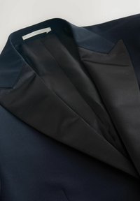 Dunkelblaues Sakko mit satinierter Revers, glatter Innenverkleidung und markanter, scharfer Kragenform. Der Stoff wirkt luxuriös und maßgeschneidert.