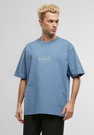 Blue cotton oversized t-shirt with short sleeves. Features a small printed graphic of a sofa and text on the front. Simple, casual design.