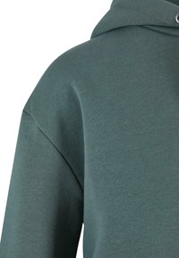 Tissu de sweat-shirt vert foncé montrant l'épaule et la capuche avec une couture visible et un œillet métallique sur la capuche.