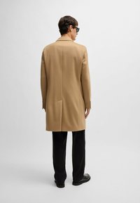Cappotto di lana beige con colletto a revers, spacco singolo e maniche con bottoni, abbinato a pantaloni e scarpe neri; design minimalista.