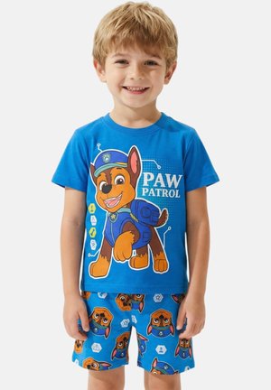 Garçon blond souriant portant un t-shirt bleu Paw Patrol et un short assorti avec des visages de personnages et des symboles de patrouille.