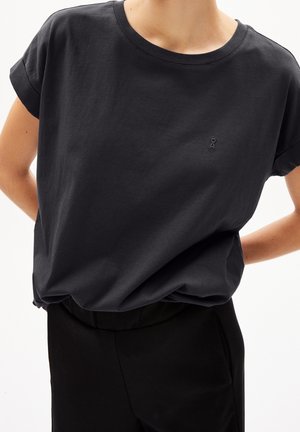 Person trägt ein locker sitzendes schwarzes T-Shirt mit kurzen Ärmeln und einem kleinen gestickten Logo auf der Brust, kombiniert mit einer schwarzen Hose.