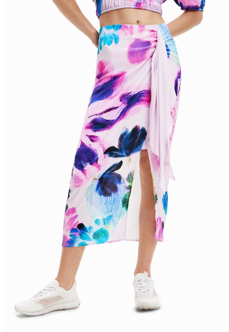 Femme portant une jupe midi à taille haute à fleurs colorées avec une fente latérale, associée à des baskets blanches et un haut court assorti.
