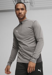 Grijze lange mouwen sportshirt met een half-rits ontwerp, voorzien van een wit logo op de borst en een gladde textuur. Draagt met zwarte broek.
