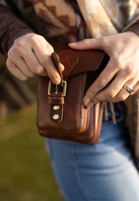 Bruin lederen zakje met metalen gesp en klinknagels, voorzien van een gestructureerd oppervlak en ronde vorm. Hand die de klep van het zakje vasthoudt.