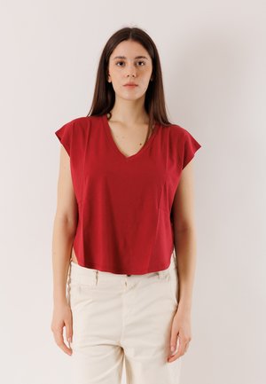 Giovane donna con lunghi capelli castani che indossa un top rosso senza maniche a scollo a V e pantaloni bianco sporco, in piedi davanti a uno sfondo bianco uniforme.