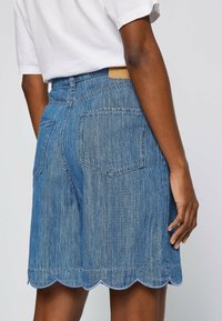 Kjol i ljusblå denim med scallop-kant, utrustad med två bakfickor och en midjebandsetikett, visad i en rak silhuett.