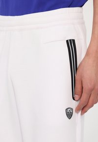Weiße Sweatpants mit schwarzen Streifen entlang der Seite, ausgestattet mit einem gestickten EA7-Logo im Bereich der vorderen Tasche und einer glatten Textur.
