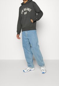 Felpa con cappuccio grigio scuro con la scritta "NEW BALANCE 1906" in bianco, tasca frontale, abbinata a jeans larghi azzurri chiari e scarpe da ginnastica argento.