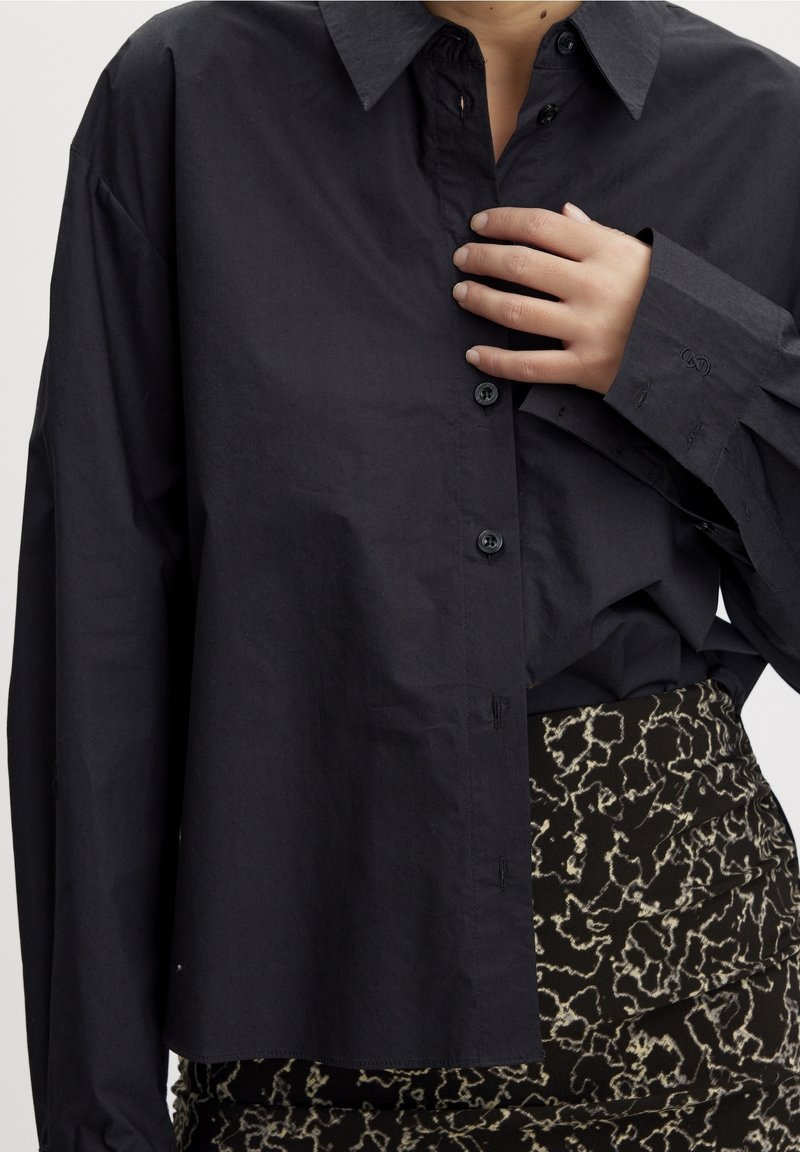 Femme portant une chemise noire oversize boutonnée et un pantalon noir et blanc à motifs, ajustant la chemise près de la poitrine avec la main.