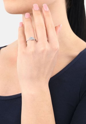 Mano sinistra con unghie manicure rosa che indossa un anello d'argento con una pietra centrale trasparente, tenuta vicino al collo e al mento di una donna.