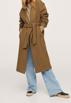 Femme portant un long manteau marron ceinturé, un jean large bleu clair, un pull beige et des chaussures à semelle épaisse noires, debout les mains dans les poches.