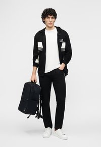 Νέος άνδρας σε ασπρόμαυρη καθημερινή στολή που κρατάει ένα μαύρο σακίδιο, στέκεται μπροστά σε απλό λευκό φόντο.