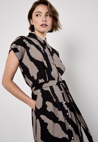 Jeune femme aux cheveux courts portant une robe à manches courtes, à motifs abstraits noirs et gris, avec une ceinture à la taille, se tenant devant un fond blanc.
