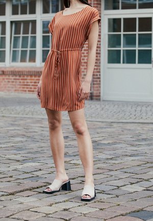 Rostfarbenes Kleid mit weißen vertikalen Streifen, tailliert mit einem Bindegürtel, kurze Ärmel, kombiniert mit beigen Slippern und schwarzen High Heels.