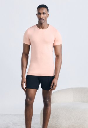 Mann steht drinnen neben einem weißen, strukturierten Sessel und trägt ein hellrosa eng anliegendes T-Shirt und schwarze Shorts.