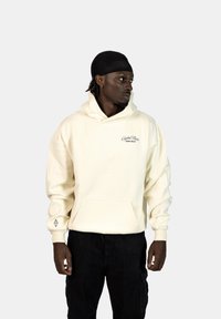 Felpa color crema realizzata in tessuto morbido, con tasca a marsupio, cappuccio con cordoncino e logo sul petto. Accentuata con pantaloni neri.