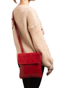 Borsa a tracolla rossa in pelle con chiusura a patta, tracolla regolabile, texture liscia e forma quadrata, indossata con un maglione lavorato a maglia beige chiaro.