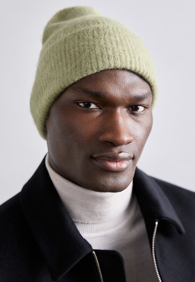 Beanie verde tricotat dintr-un material moale, cu o textură înclăcată. Purtați cu un guler înalt deschis la culoare și o jachetă închisă, pe un fundal neutru.
