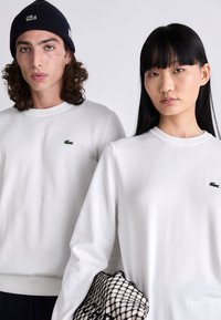 White cotton sweaters with ribbed cuffs and crew necks, featuring a small green Lacoste logo on the left chest. Black knit beanie worn.