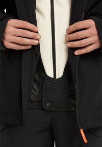 Zwarte buitenjas met een witte fleece binnenvoering, voorzien van een ritssluiting aan de voorkant, een drukknoopsluiting bij de taille en een oranje ritsaccent.