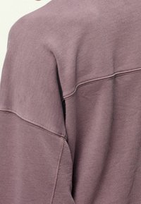 Světlofialová mikina z texturované, měkké látky. Má spadaná ramena a viditelné švy podél švů.