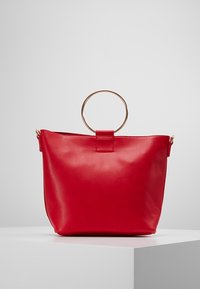 Rød syntetisk håndtaske med en skinnende gylden cirkulær hank, blød tekstur og enkelt design. Har lukning foroven og ingen yderligere mønstre.