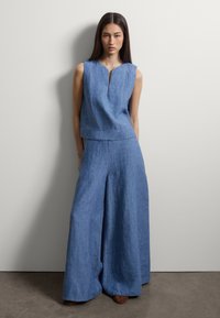 Mujer con una blusa sin mangas de lino azul con escote en forma de muesca y pantalones anchos a juego, de pie frente a una pared lisa y clara.