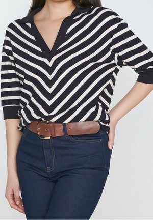 Femme portant un haut rayé noir et blanc à col en V, un jean bleu foncé et une ceinture en cuir marron, avec une main posée sur la hanche.