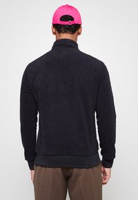 Giacca in pile blu navy con collo alto, polsini e orlo a costine. Texture liscia; indossata con un cappellino rosa acceso. Vista posteriore solo.