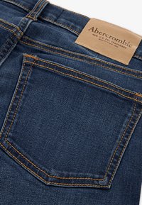 Σκούρες μπλε τζιν παντελόνες με πίσω τσέπη και πορτοκαλί ραφή, καθώς και μπεζ δερμάτινη ετικέτα μάρκας Abercrombie πάνω από την τσέπη.